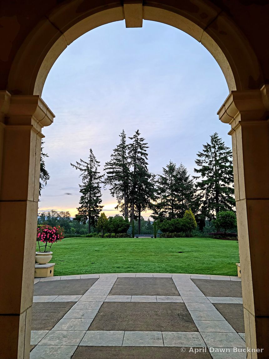 Looking out from under a stone archway to a paved patio and green lawn, with tall trees beyond