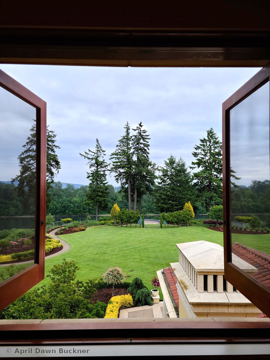 Through open windows to a stone terrace, the lawn and evergreens under a cloudy sky
