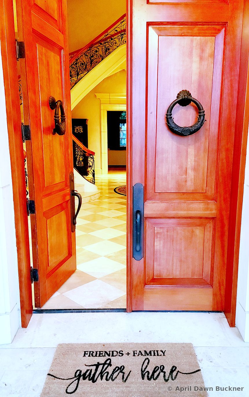 Open wooden front doors with round metal knockers and a “FRIENDS + FAMILY gather here” doormat, with a tiled floor and the foot of the stairs inside