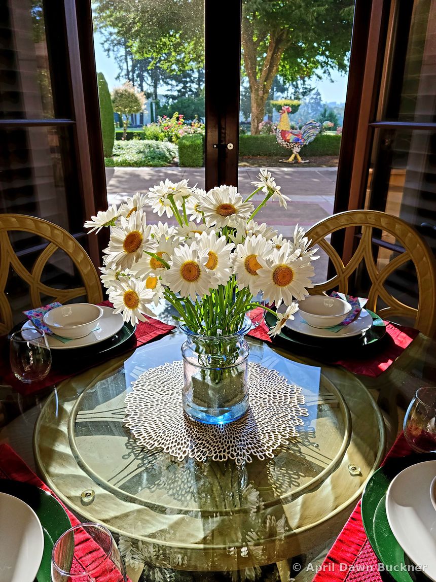 A round glass table set for a meal with white plates, red napkins and a vase of white daisies