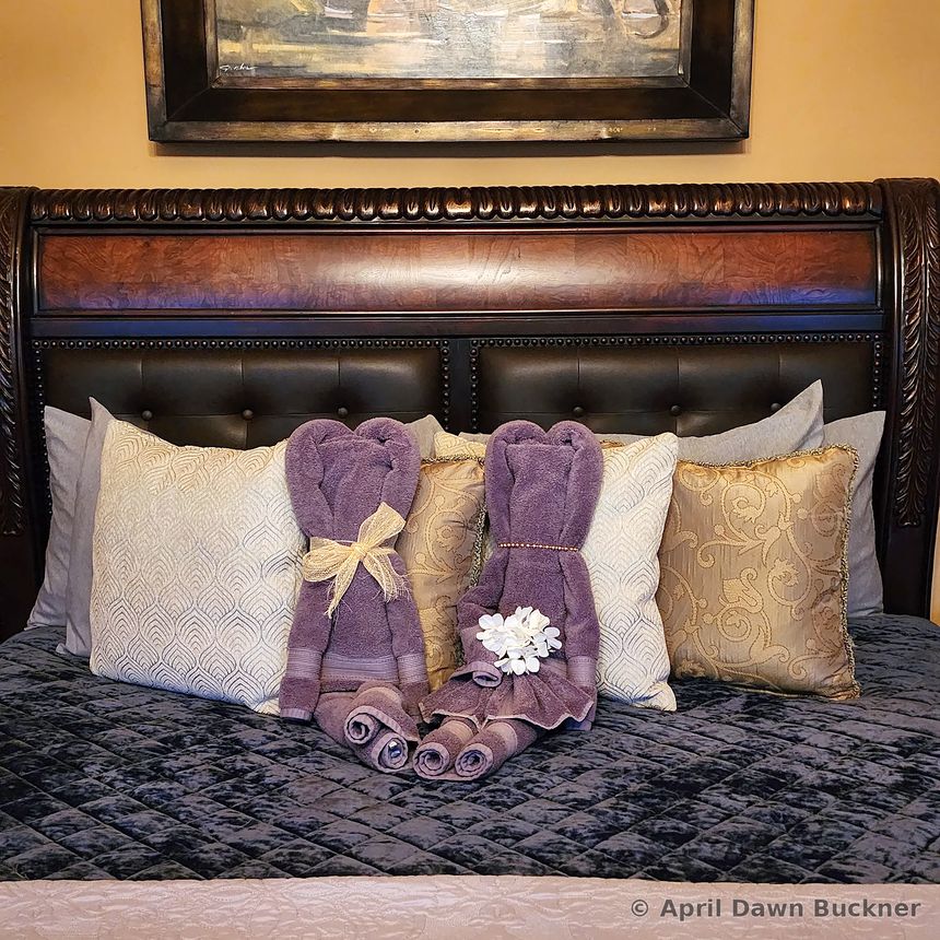 A bed with a tufted dark headboard, white pillows and two purple towels folded into decorative shapes