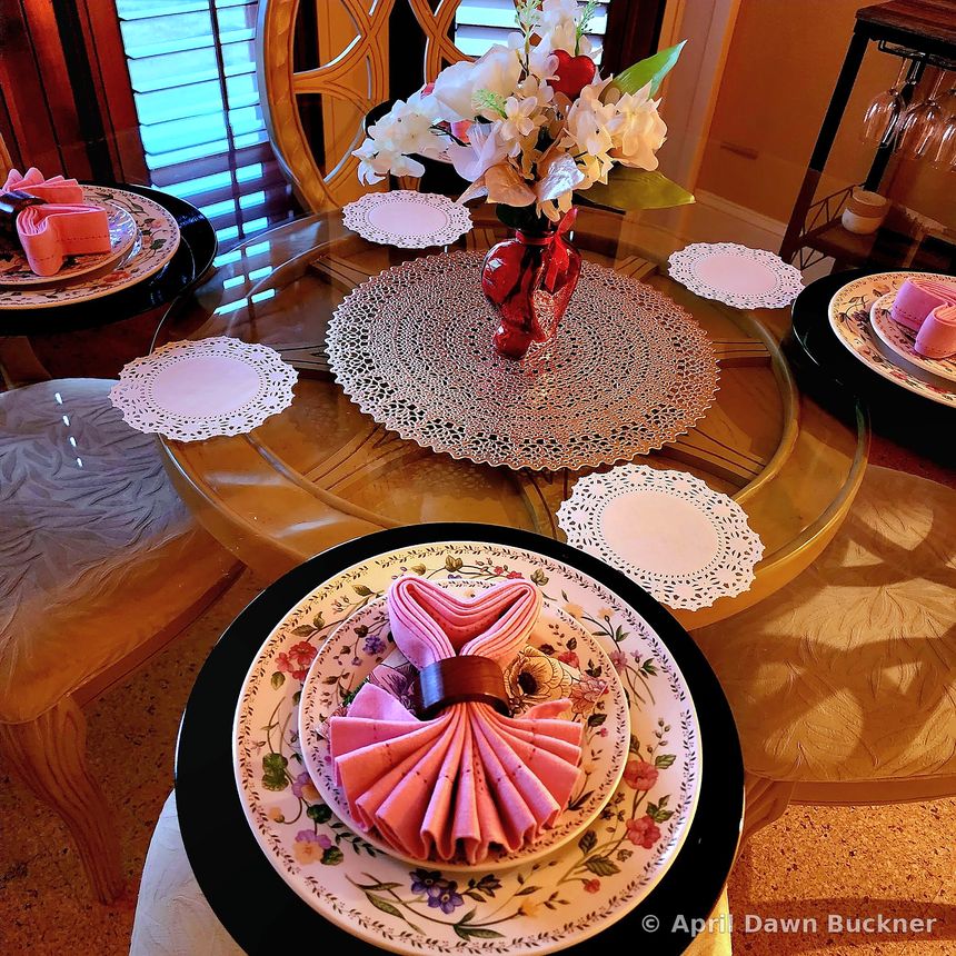 A round glass table set for a meal, seen from above, with floral plates, gold placemats, pink napkins and a vase of flowers