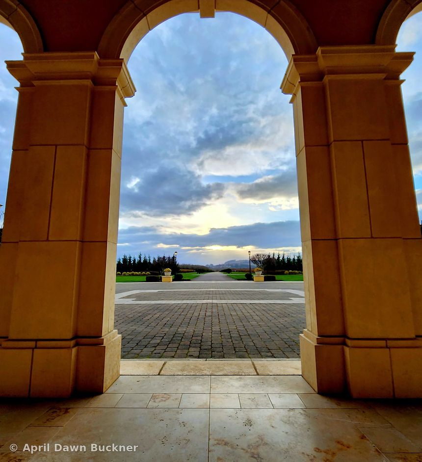 Through a large stone archway to the paved courtyard, lawn and trees at sunset