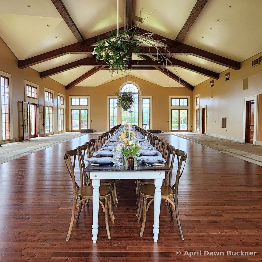 The large beamed hall with a long banquet table set, greenery hanging from the ceiling beams, and light from big windows