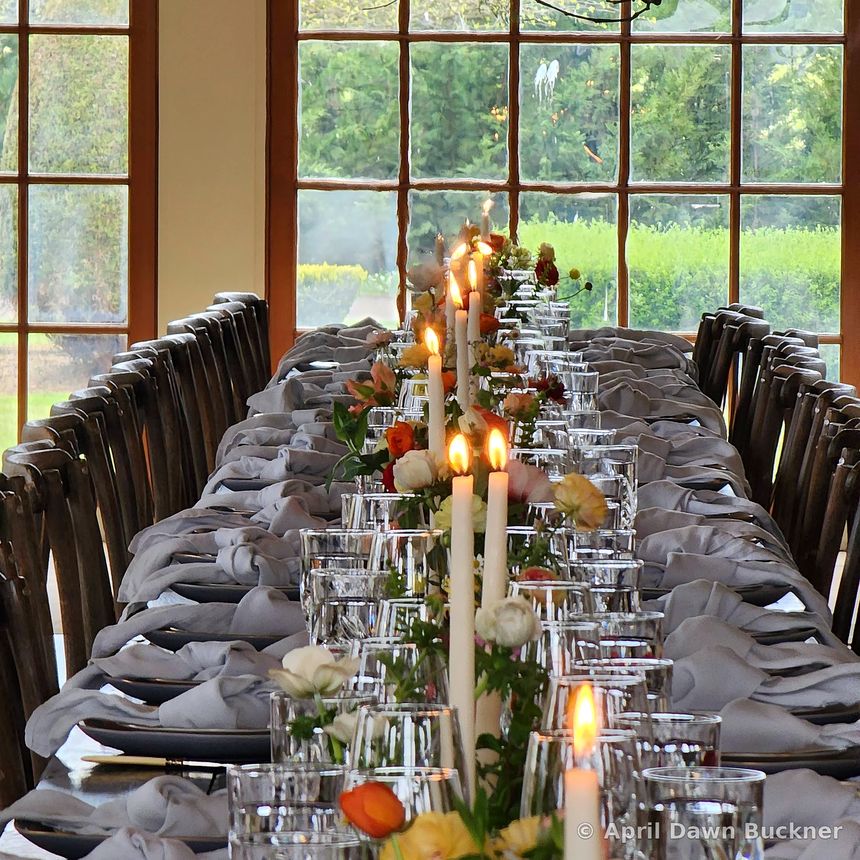 A set table with tall white candles, glassware and flowers, windows behind