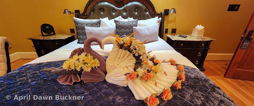 A bed with two towels folded as swans and orange flowers, with dark wood nightstands and lamps