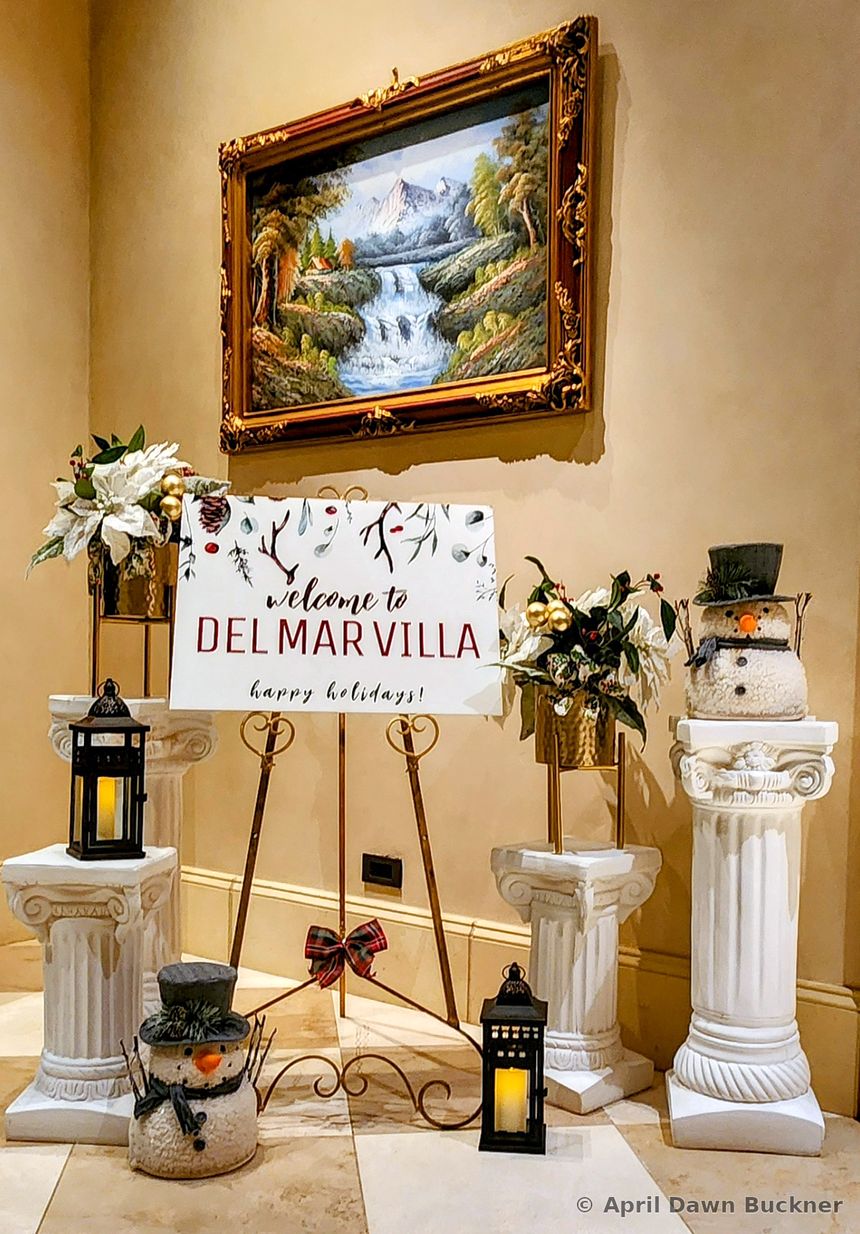 A “Welcome to Del Mar Villa – Happy Holidays!” sign on an easel with greenery, lanterns and snowman figures, and a landscape painting behind