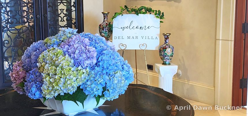 A large bouquet of blue and purple hydrangeas with a small “Welcome to Del Mar Villa” sign behind it