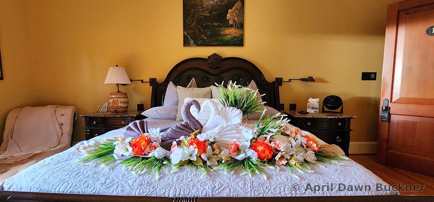 A bed with two white towel swans forming a heart, surrounded by orange and white flowers