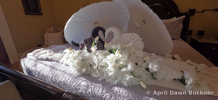 A bed with white towel swans, white flower garlands and two open white paper parasols