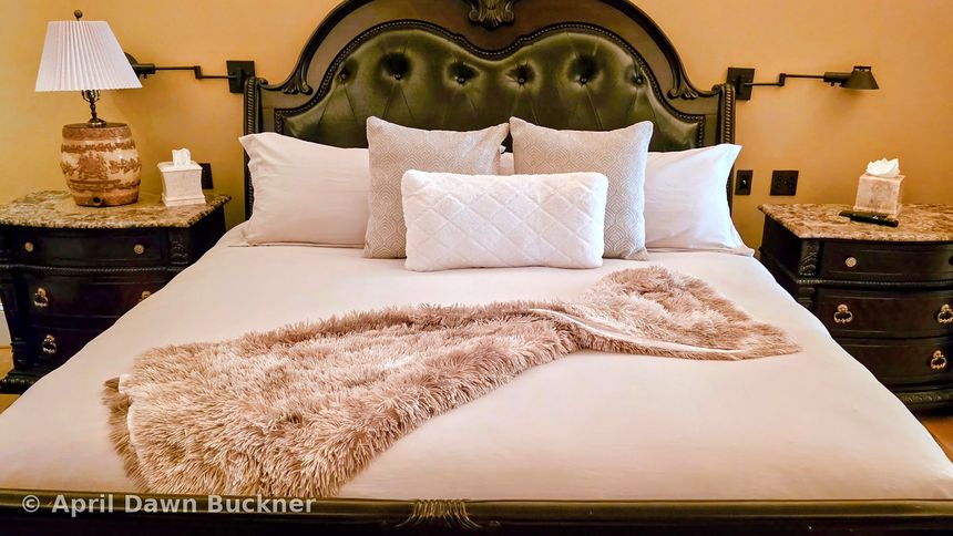 A bed with a dark green tufted headboard, white linens and a tan faux-fur throw